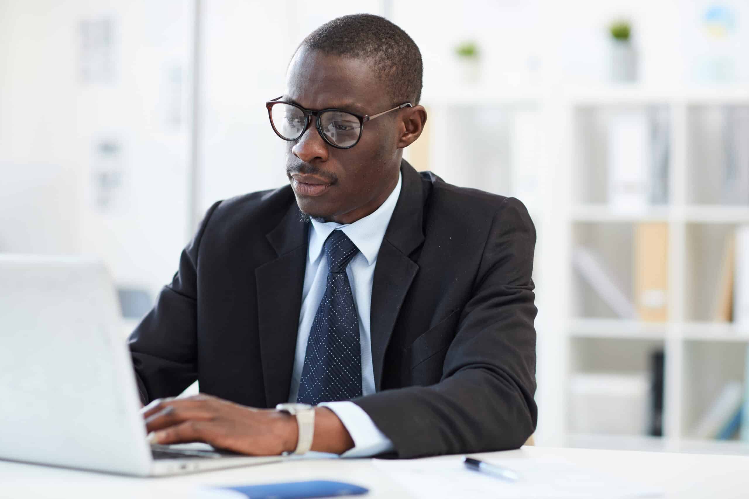 Concentrated young African businessman wearing eyeglasses and formal wear working on laptop computer at the workplace