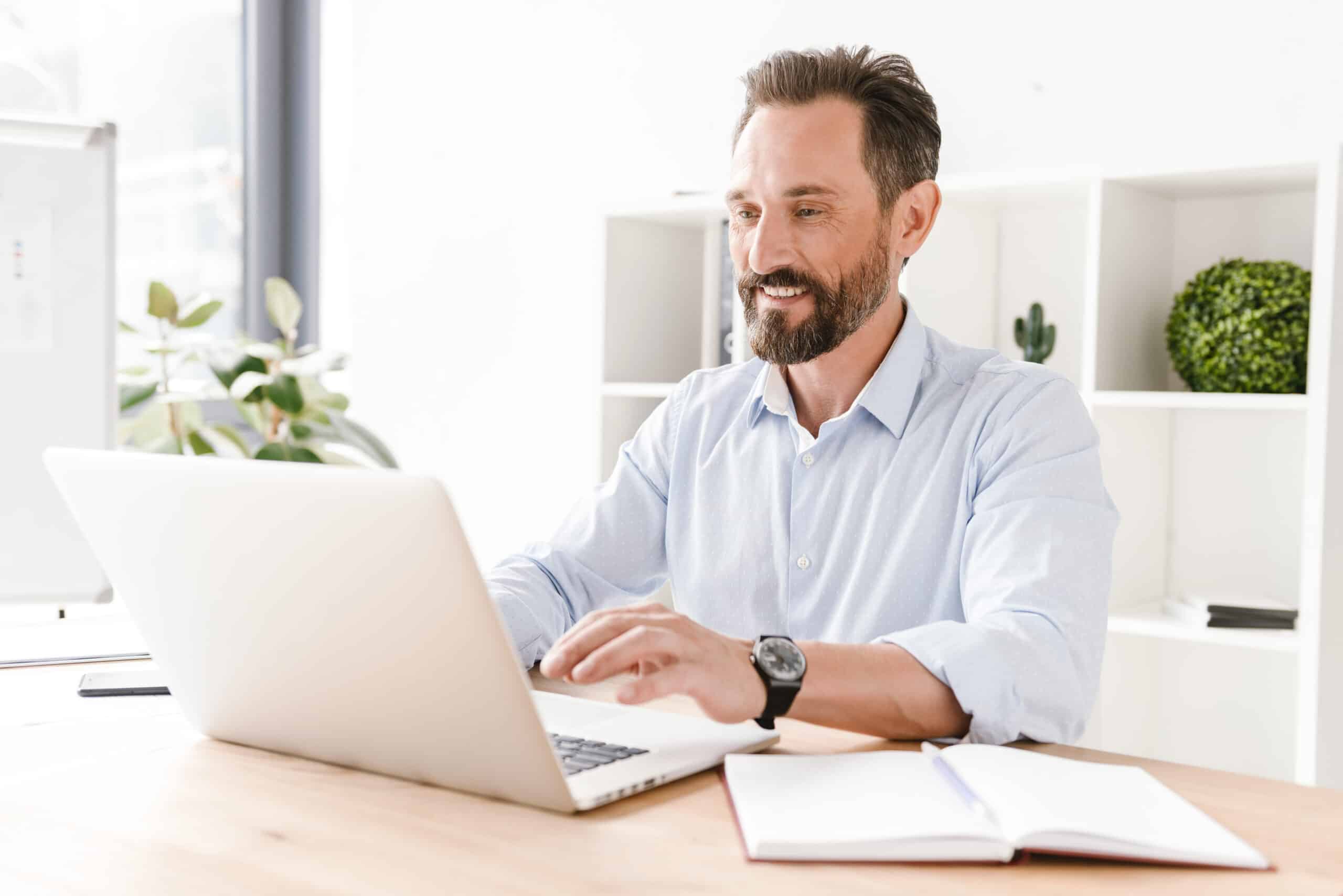 Smiling businessman working on laptop computer while sitting at the office desk with paperwork